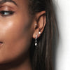 A model wearing Juraster’s Sterling Silver Star Earring Charm, Star-Drop, with an engraved star, crafted from Fairmined sterling silver, attached to a white gold pavé diamond hoop, a sterling silver pearl stud in her second and a white gold diamond stud in her tragus.