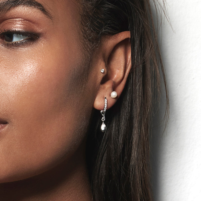 A model wearing Juraster’s Sterling Silver Star Earring Charm, Star-Drop, with an engraved star, crafted from Fairmined sterling silver, attached to a white gold pavé diamond hoop, a sterling silver pearl stud in her second and a white gold diamond stud in her tragus.